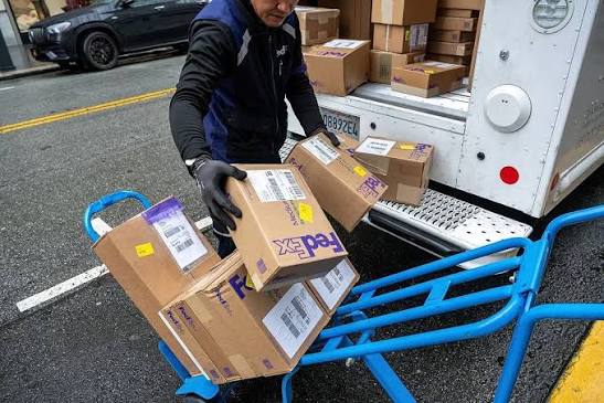 FedEx courier loading cardboard boxes onto a hand truck next to a delivery van